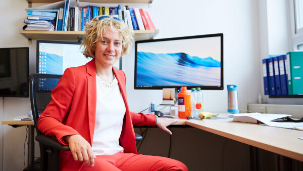 Стефани Вехнер обращается к камере, на заднем плане на её столе стоят два компьютерных монитора.