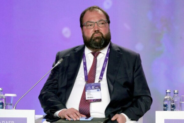 Человек в деловом костюме на конференции, сидит за столом с микрофоном и бутылками воды.