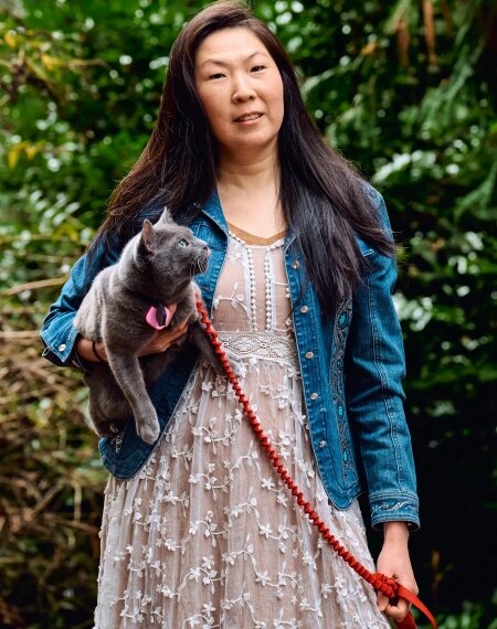 Женщина на прогулке в джинсовой куртке держит серого кота на поводке в саду.