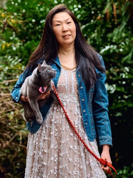 Женщина на прогулке в джинсовой куртке держит серого кота на поводке в саду.