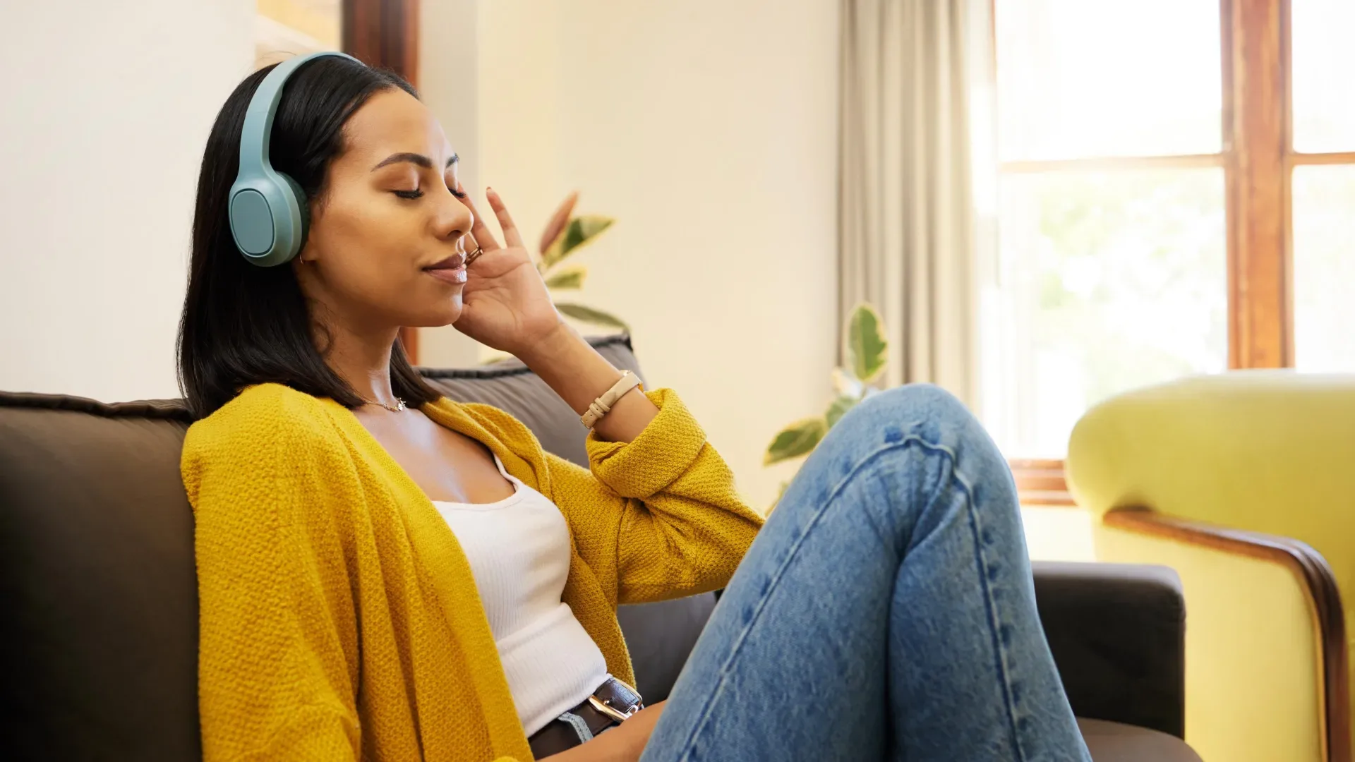 Человек в наушниках расслабляется на диване, одет в жёлтый кардиган и джинсы.