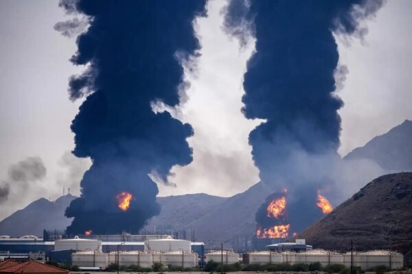 Пожар на нефтеперерабатывающем заводе, дым и огонь на фоне горного пейзажа.