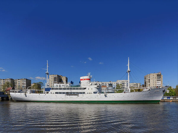 Белое судно у берега с многоэтажками на фоне, солнечный день.