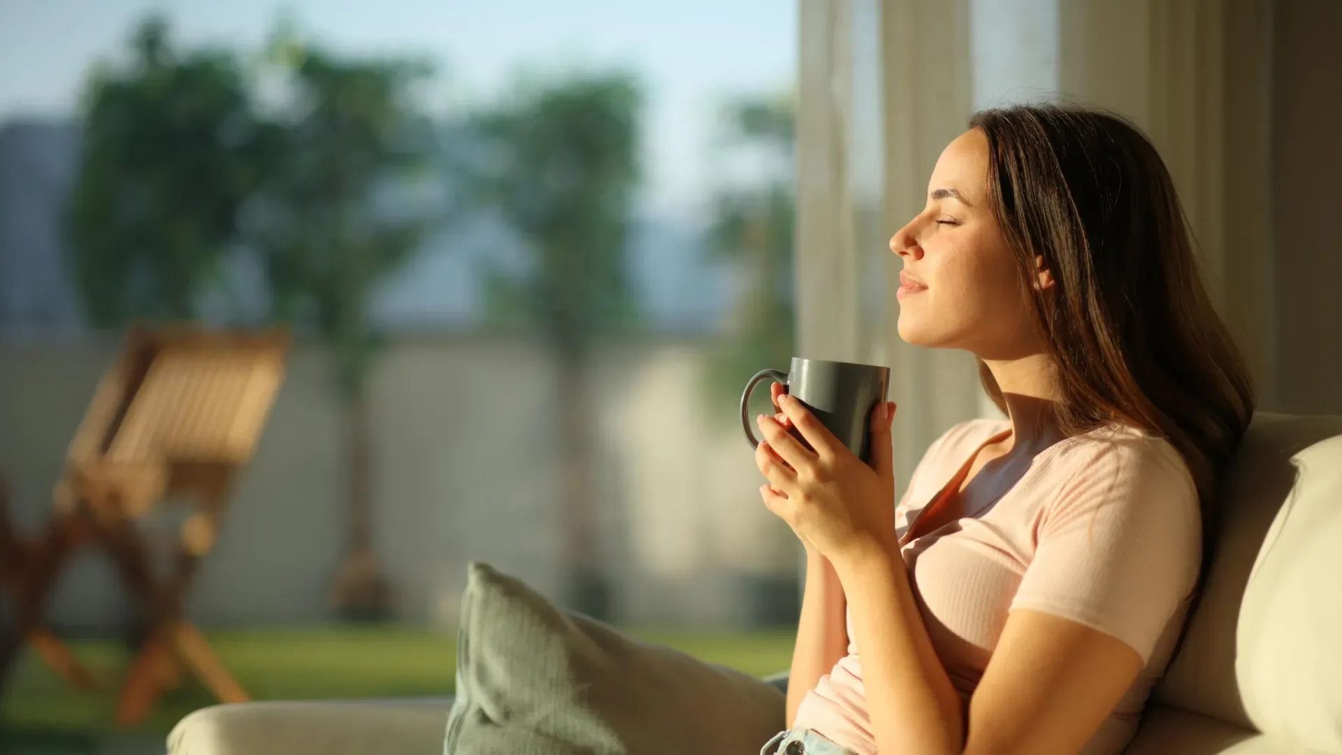 Женщина с чашкой чая сидит на диване, наслаждаясь утренним солнцем в гостиной.