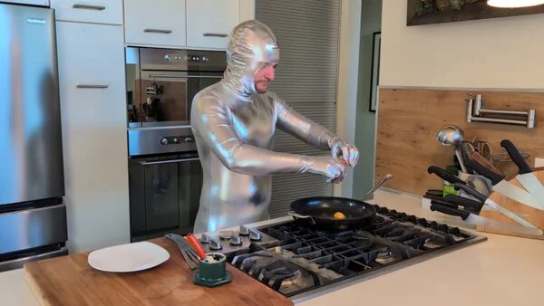 Человек в серебряном костюме готовит яичницу на кухне.