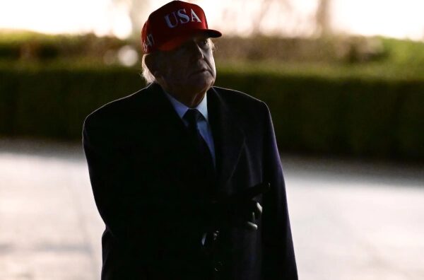 Изображение человека в красной кепке с надписью USA в темноте, на улице.