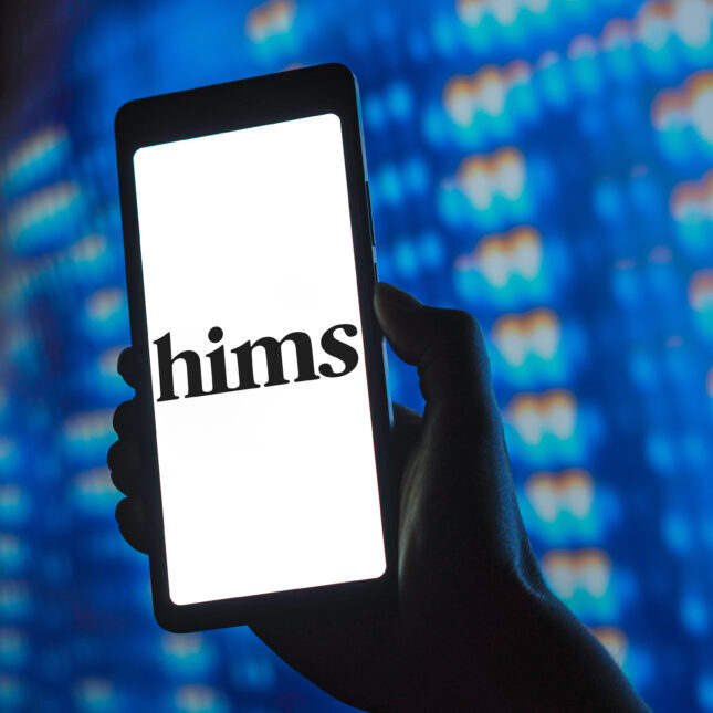 Смартфон с логотипом "hims" на экране, в руке на фоне синих огней.