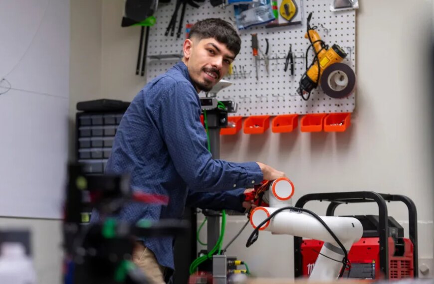Мужчина работает с роботом в мастерской, окружён инструментами на фоне.