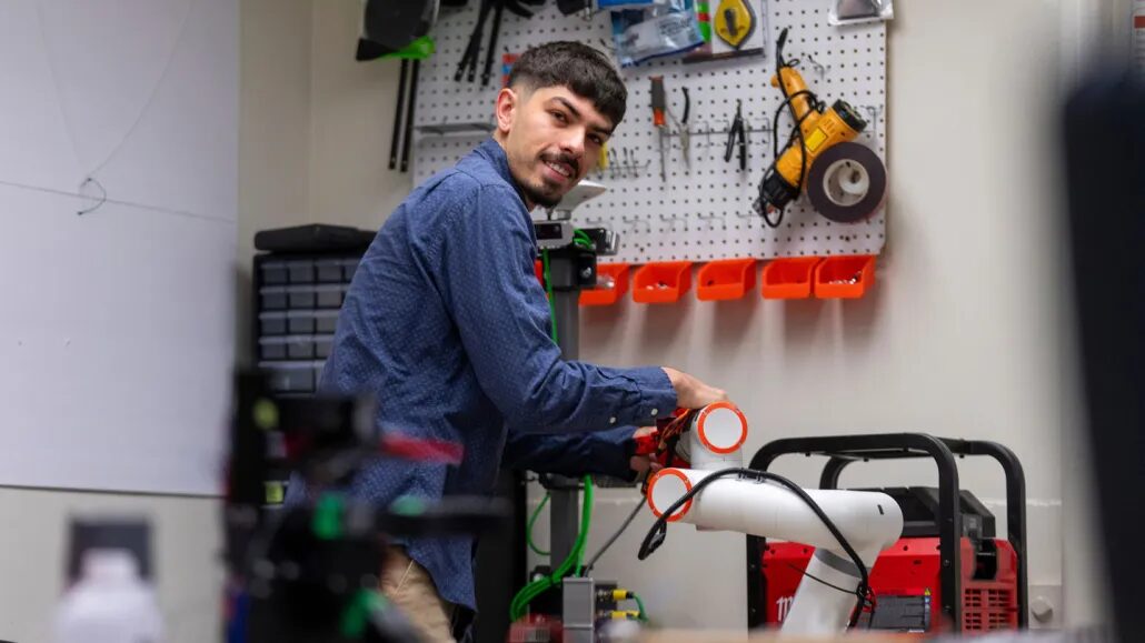 Мужчина работает с роботом в мастерской, окружён инструментами на фоне.