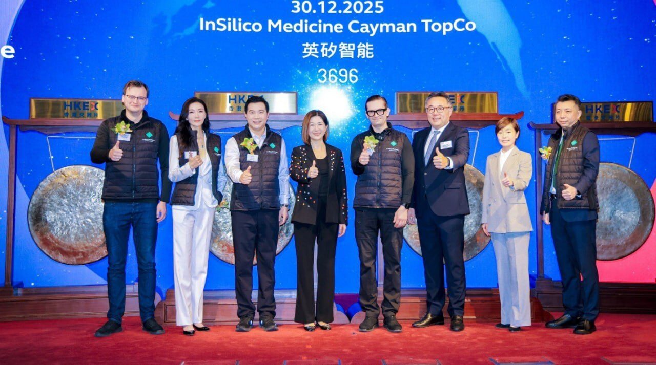 Группа людей в костюмах и жилетах улыбается и показывает жест "класс" на фоне баннера.