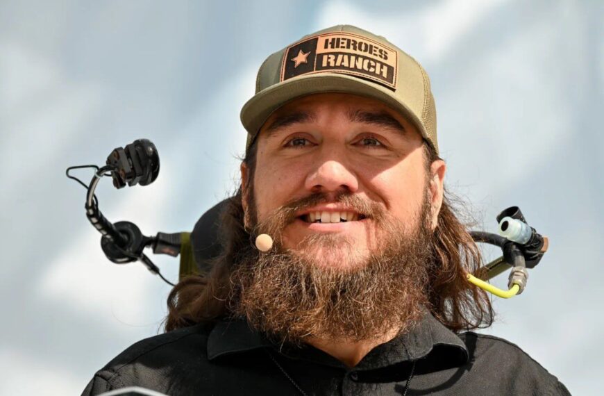 Мужчина в бежевой кепке с микрофоном на фоне неба.