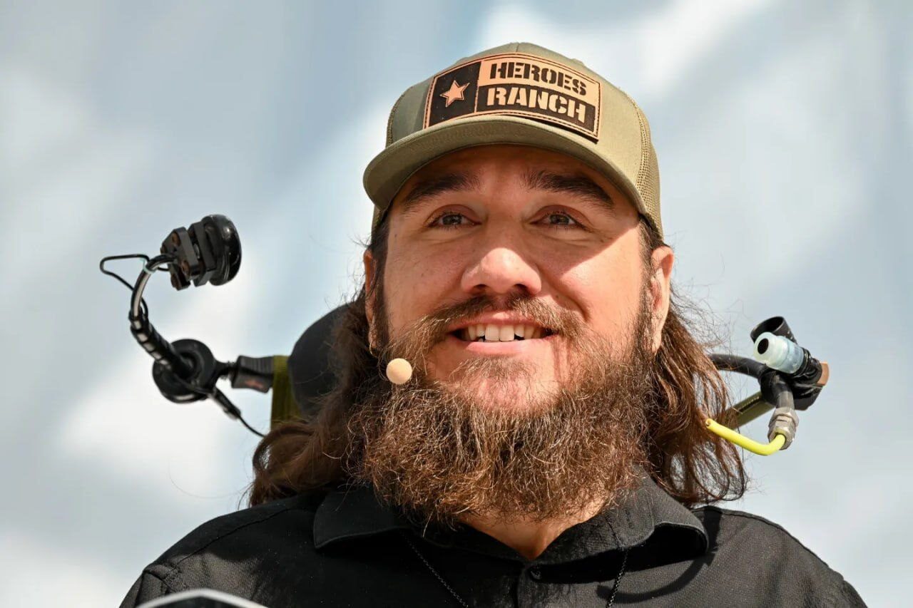 Мужчина в бежевой кепке с микрофоном на фоне неба.