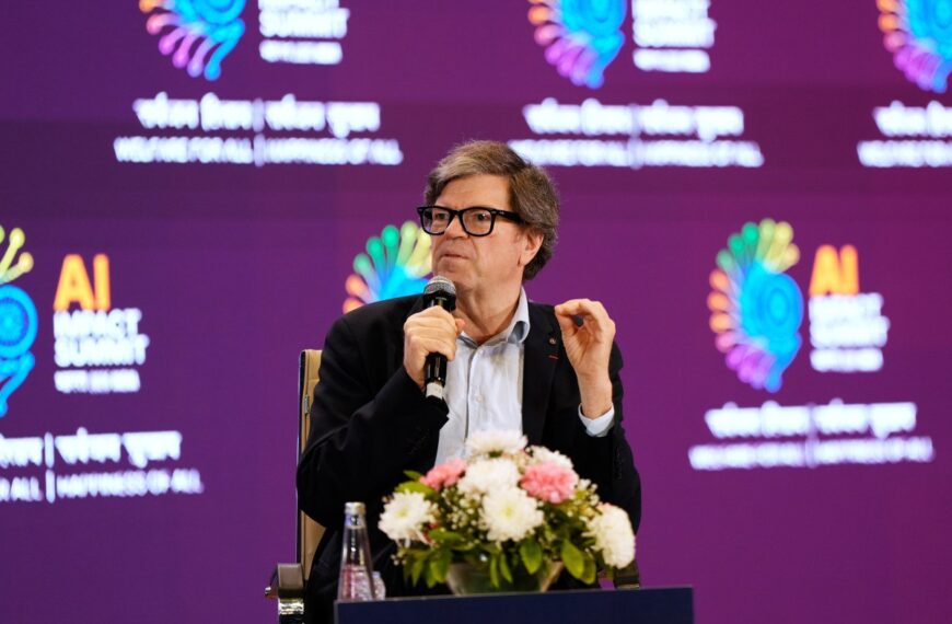 Спикер с микрофоном на AI Impact Summit, цветы на столе, яркий фон.