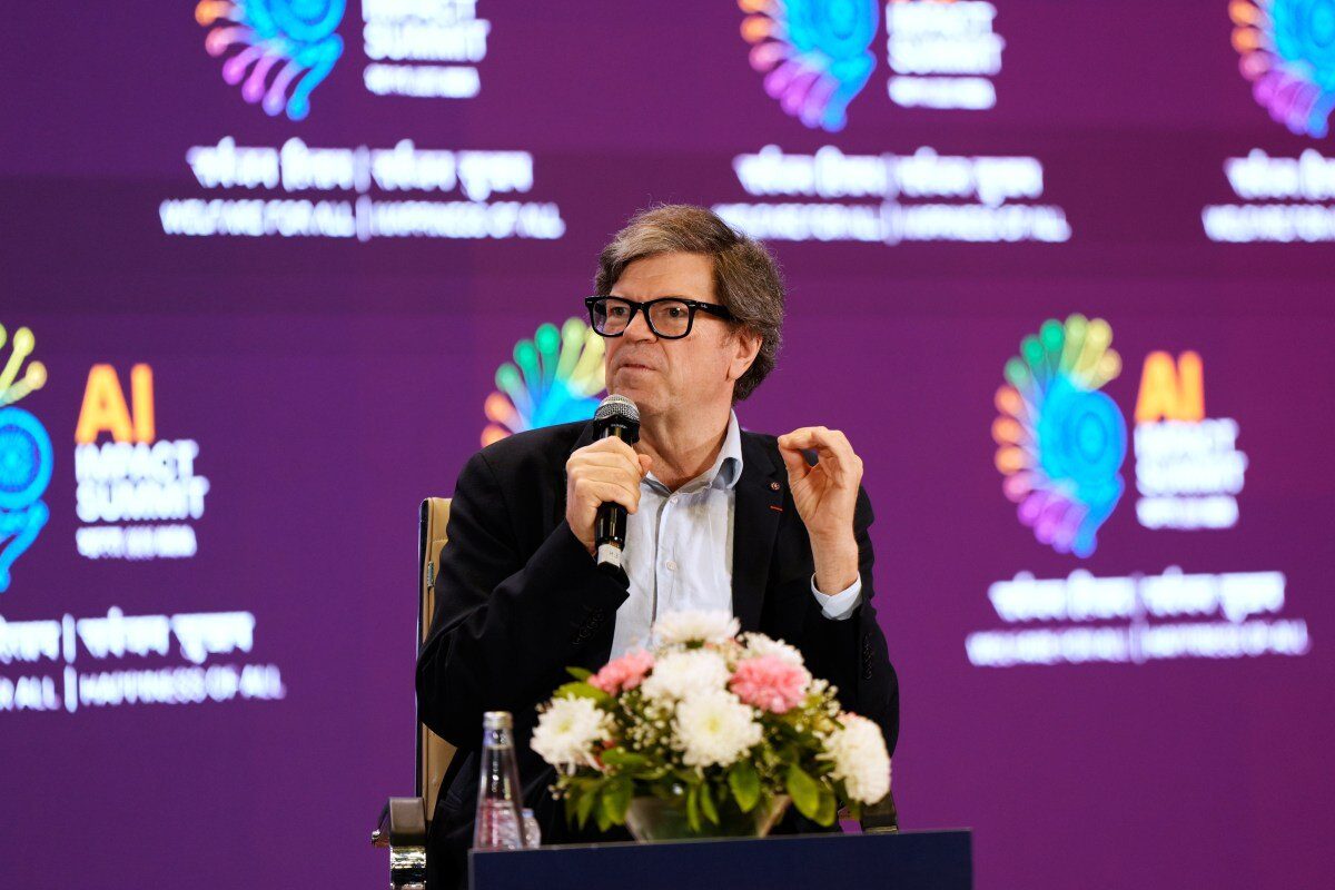 Спикер с микрофоном на AI Impact Summit, цветы на столе, яркий фон.