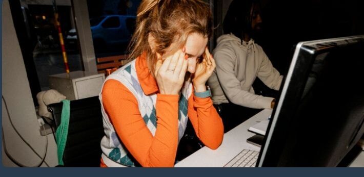 Женщина в офисе сидит за компьютером, закрывая лицо ладонями.