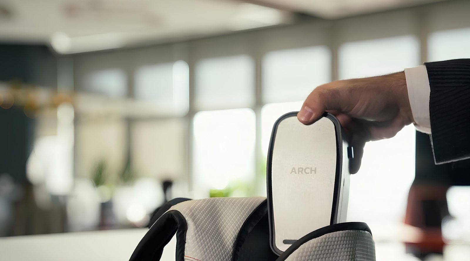 Рука в пиджаке кладет устройство ARCH в рюкзак на фоне офиса.
