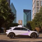 Автономный автомобиль Avride в городском пейзаже, окружённый деревьями и высотками.