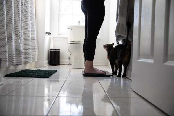 Ведущий эксперт Виктория Сонг стоит на весах в ванной комнате, а кошка наблюдает за ней.