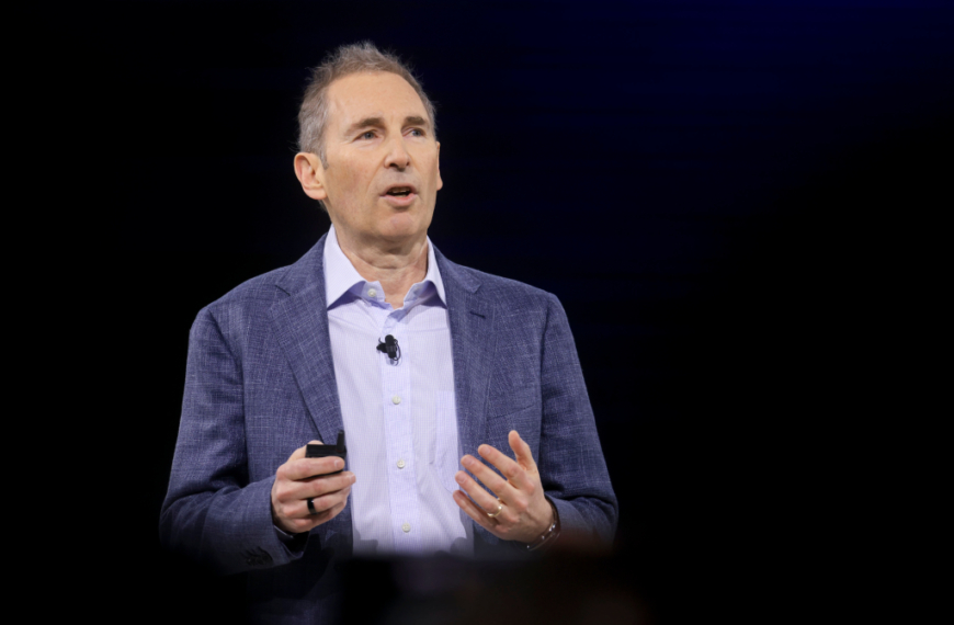 Человек в пиджаке и рубашке выступает с речью на темном фоне.