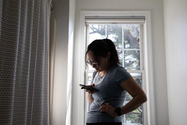 Ведущий эксперт Виктория Сонг рассматривает приложение для непрерывного мониторинга уровня глюкозы на телефоне в ванной комнате, стоя перед окном.