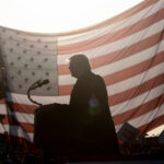 Силуэт человека у микрофона на фоне флага США, освещённого солнцем.
