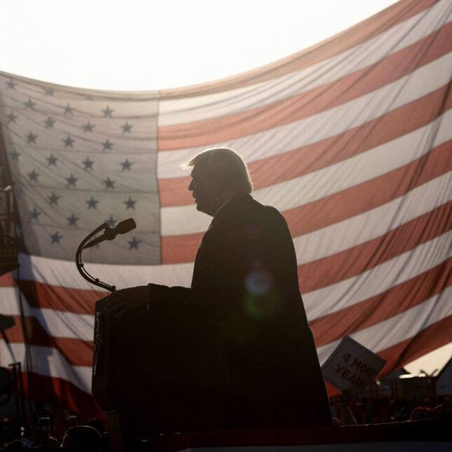 Силуэт человека у микрофона на фоне флага США, освещённого солнцем.