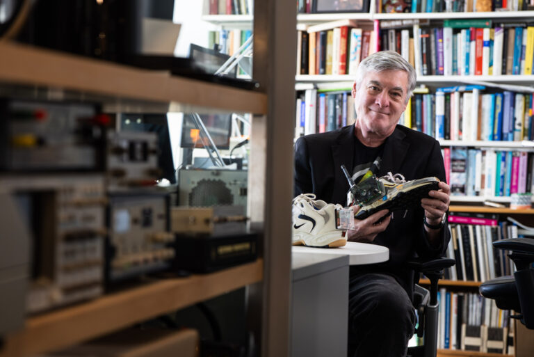 Человек изучает кроссовку с электроникой в офисе с книгами на заднем плане.