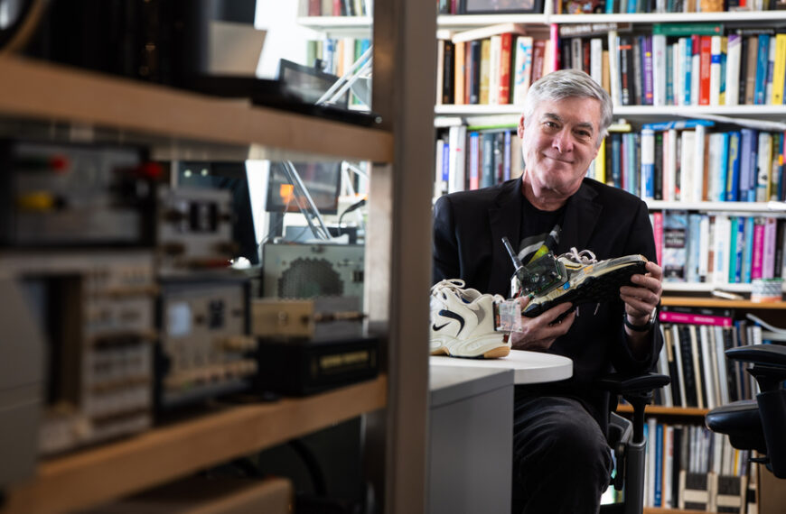 Человек изучает кроссовку с электроникой в офисе с книгами на заднем плане.