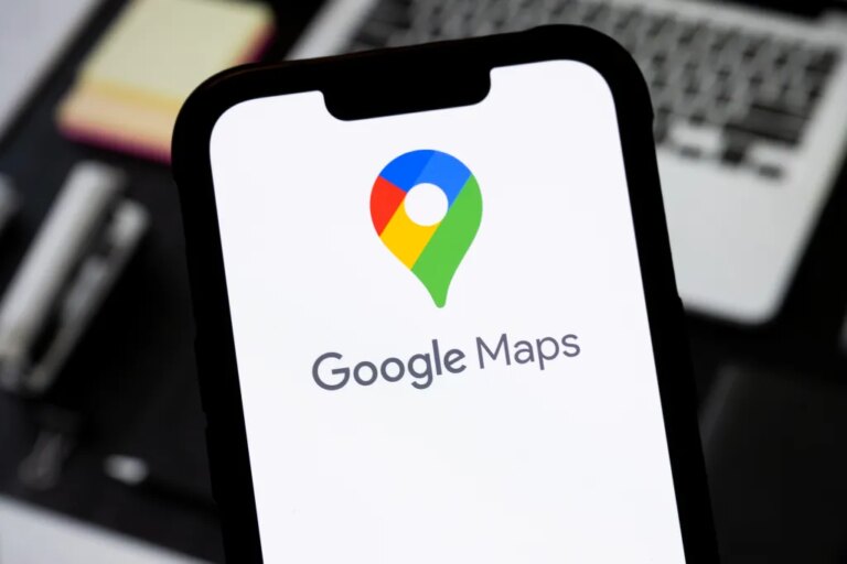 Смартфон с логотипом Google Maps на экране, фон с ноутбуком и блокнотом.