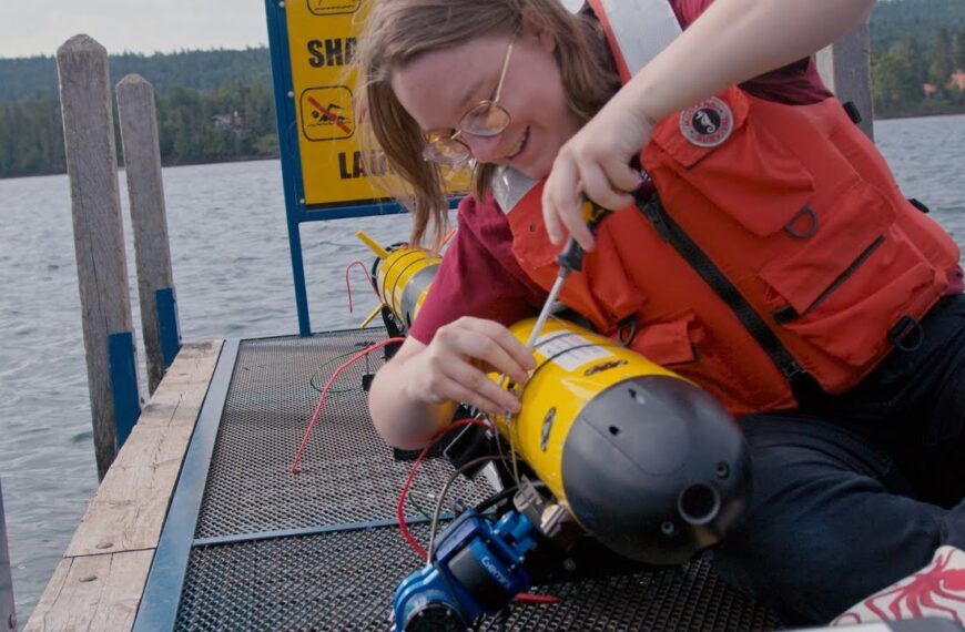 Человек в красном жилете готовит подводный дрон на причале у озера.