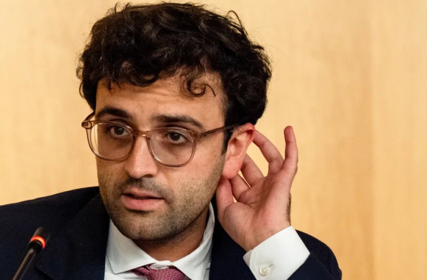 Человек в костюме и очках прислушивается, поднеся руку к уху.