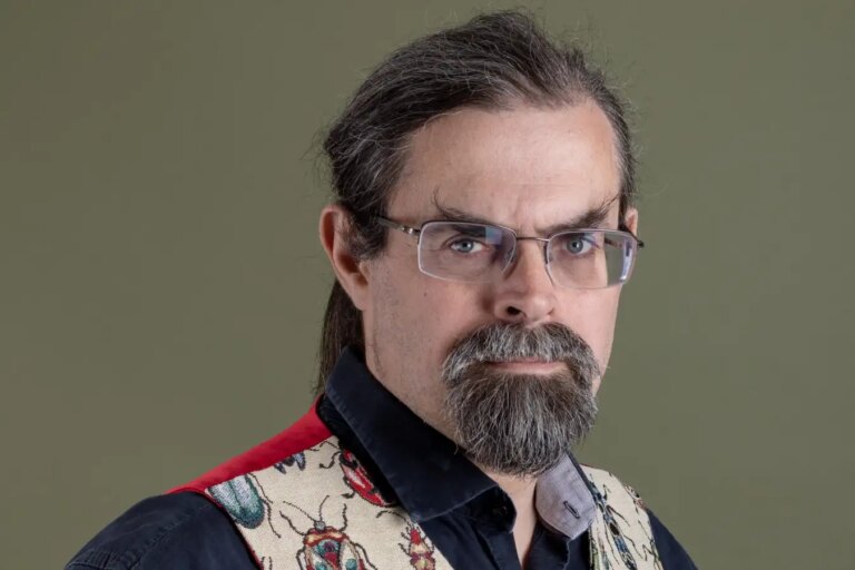 Человек в очках и жилете с жуками на фоне нейтрального цвета.