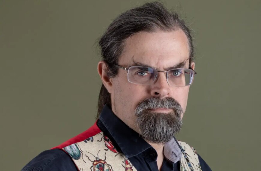 Человек в очках и жилете с жуками на фоне нейтрального цвета.