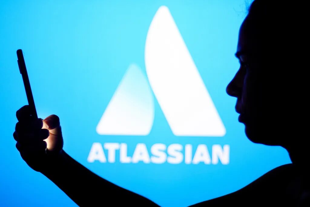 Компания Atlassian запускает инструменты визуального искусственного интеллекта и сторонних агентов в Confluence. 1 Логотип Atlassian на синем фоне, на переднем плане силуэт человека, держащего телефон, по какой-то причине.