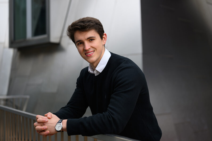 Мужчина в черном свитере улыбается, опираясь на перила возле современного здания.