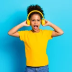 Ребенок в желтой футболке слушает музыку в наушниках на синем фоне.