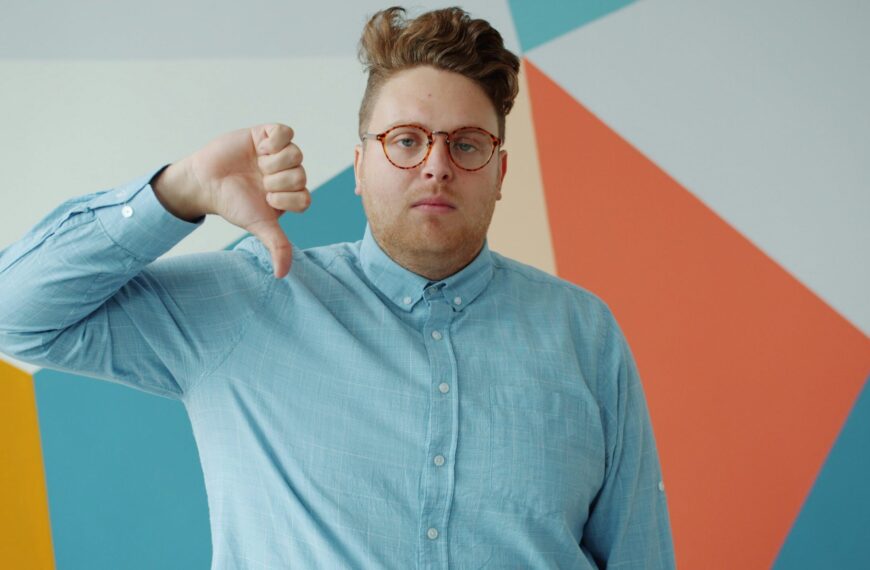 Мужчина в голубой рубашке показывает жест "палец вниз" на фоне цветной стены.