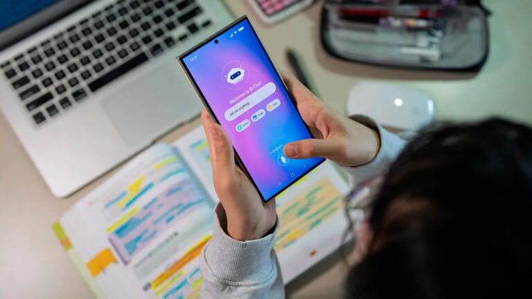 Человек держит смартфон с открытым AI-чатом, ноутбук и блокнот на столе.