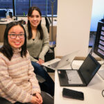 Две женщины в офисе обсуждают графики на экране, сидя за компьютерами.