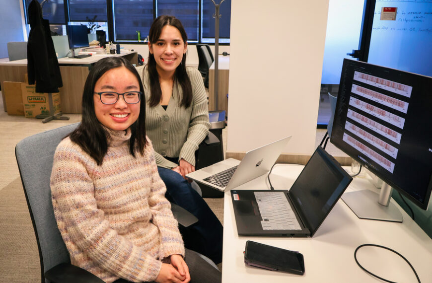 Две женщины в офисе обсуждают графики на экране, сидя за компьютерами.