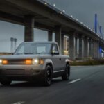 Электропикап Rivian R1T едет по дороге под мостом на фоне вечернего неба.
