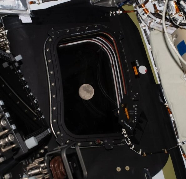 Луна видна через иллюминатор космического корабля, окруженного проводами и оборудованием.