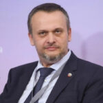 Мужчина в костюме на фоне стены с логотипом и текстом, микрофон сбоку.