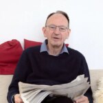 Мужчина на диване читает газету, окружён красными подушками и светлым пледом.