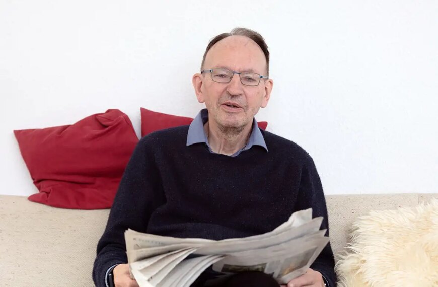 Мужчина на диване читает газету, окружён красными подушками и светлым пледом.