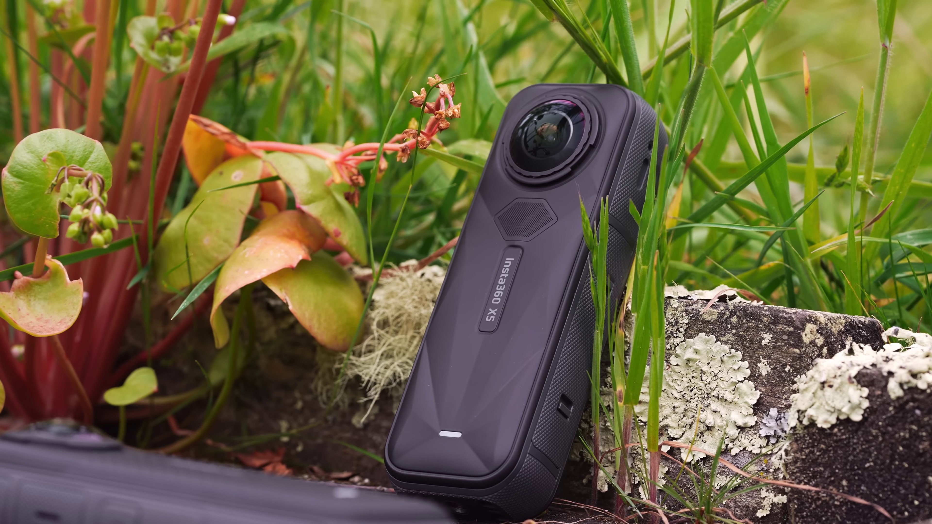 Черная 360-градусная камера-палка Insta360 X5, установленная на клумбе с цветами и травой.