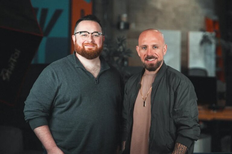 Два человека стоят рядом в помещении, один в серой кофте, другой в темной куртке с татуировками.