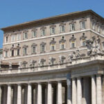 Дворец в Ватикане с колоннами и статуями на фасаде на фоне синего неба.