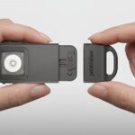 Руки держат устройство Pebblebee с открытым отсеком; серый корпус и белая внутренняя часть.