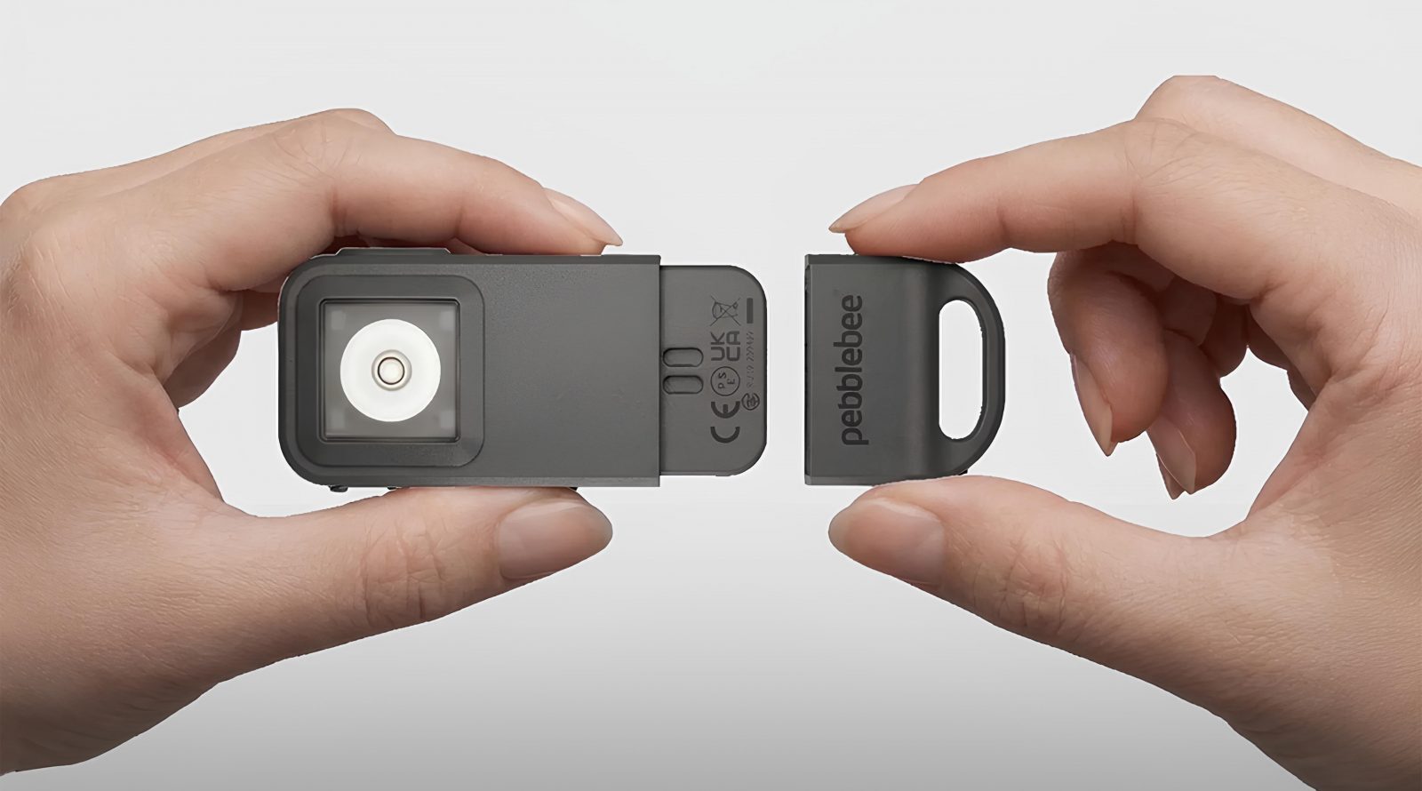 Руки держат устройство Pebblebee с открытым отсеком; серый корпус и белая внутренняя часть.